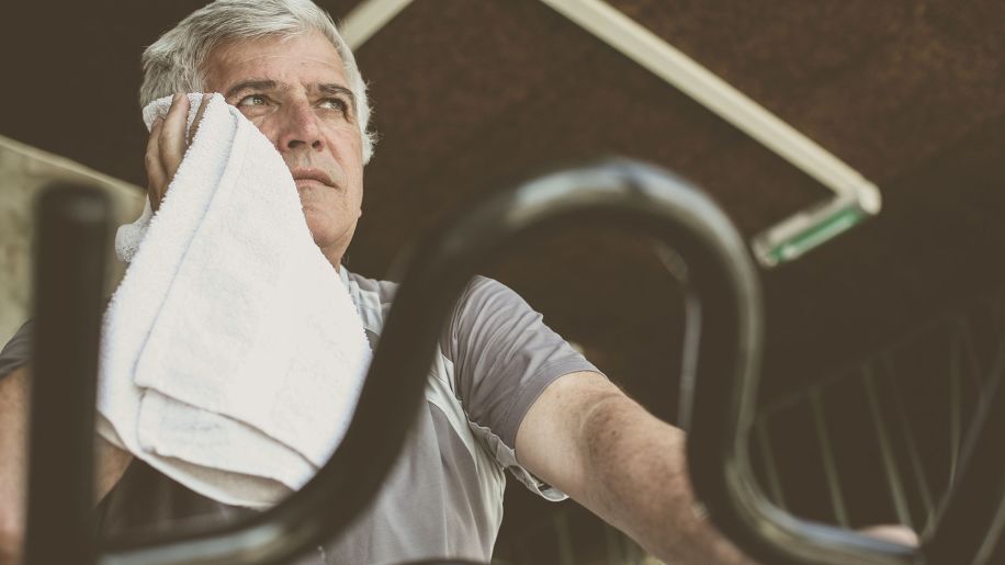 Middle-aged man wipes away sweat with a towel while working out in a gym.