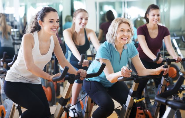 Positive happy females of different age training on exercise bikes together in fitness club
