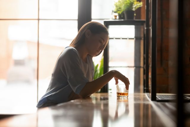 A woman with signs of alcohol addiction sits at a bar with a drink in the middle of the day. She has addiction warning signs.