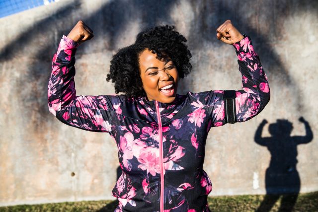 A Black woman in exercise clothes raises her arms in celebration after a workout.