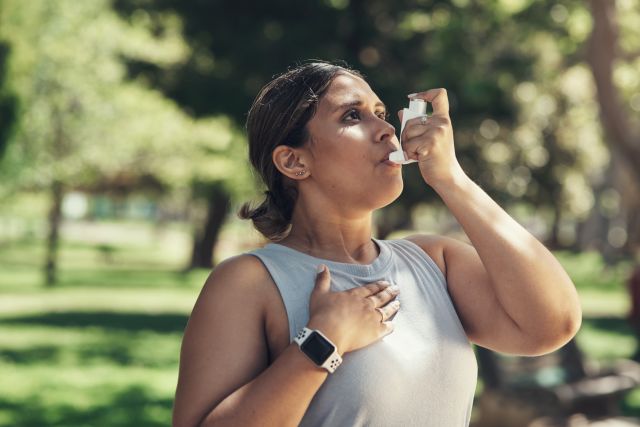 a Latino woman standing outside on a sunny day uses an asthma inhaler 