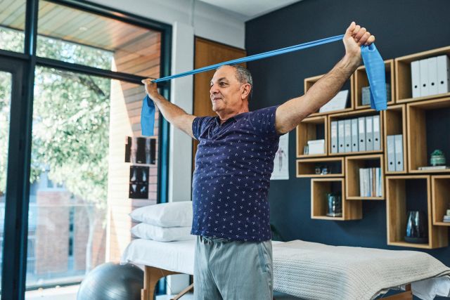Middle-aged man looking positive and strong while he strengthens his muscles and bones by stretching a resistance band above his head