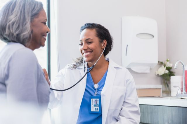 Young Black healthcare provider smiling and using a stethoscope to listen to the heart of a middle-aged Black woman, who is also smiling