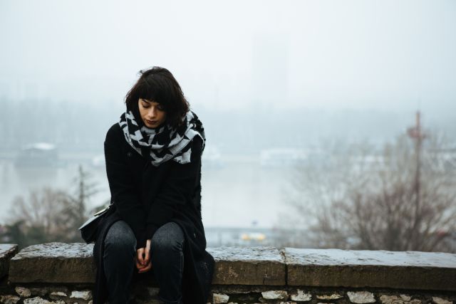 a young woman with a downcast expression sits on a stone wall on a cold, dark, winter day