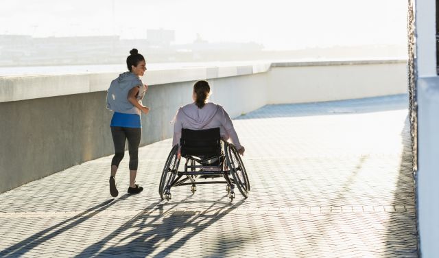 A woman in a wheelchair exercises with her friend along a city waterfront.