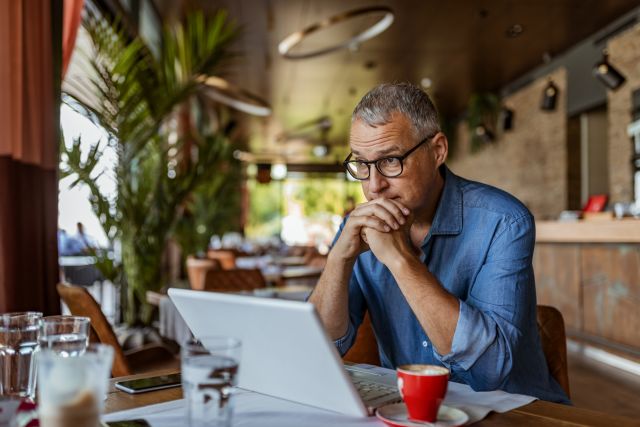 a middle aged white man sits at his computer, appearing to be fatigued or frustrated