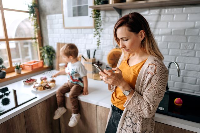 young blonde mother staring at smartphone distracted, child beside her on kitchen counter alone