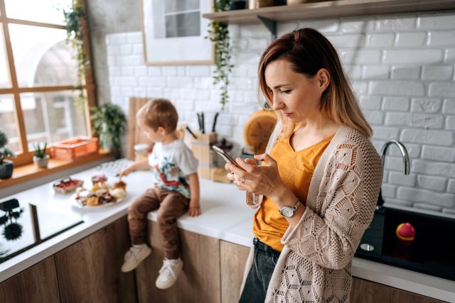 young parent staring at smartphone distracted, child beside them on kitchen counter alone