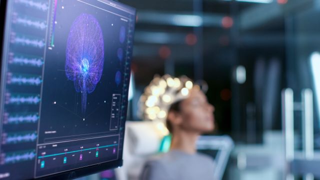 closeup of eeg reading on a monitor with a woman in the background wearing a brain scanning headset