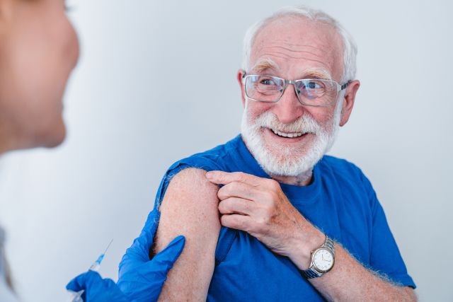 An older white man smiles as he is about to receive a vaccination at a healthcare provider's office.