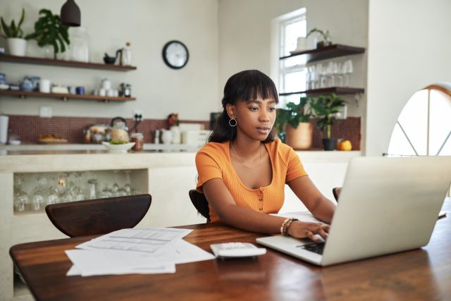 Young woman sits at her kitchen table looking over her finances on a laptop