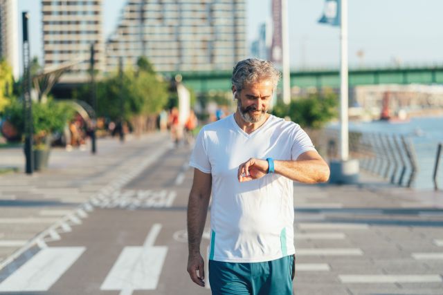 Mature man walks on a boardwalk wearing wireless headphones looking at his pedometer watch for a step count