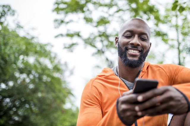 African american man sitting outdoors before his training. He is holding mobile phone and looking at it.