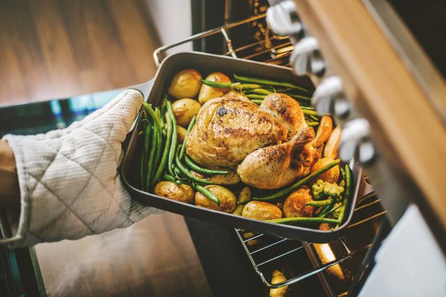 Hand wearing over mitt taking pan of baked chicken with vegetables from the oven.