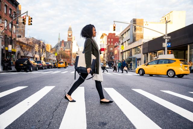 smiling woman in her 30s walking in a busy city crossing the street holding coffee 