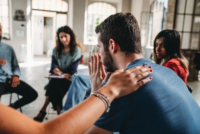 View of an addiction support meeting, a group of people and a drug counselor sit in a circle, a woman's hand resting on a man's shoulder while he shares