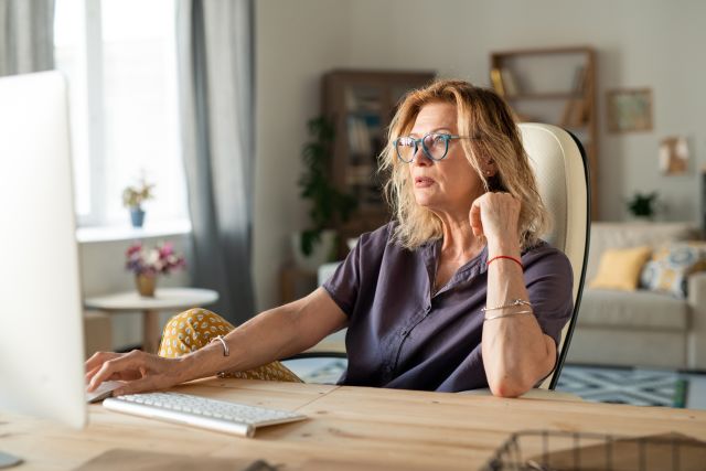 a middle aged white woman working from her office sits at a desk in front of her computer 