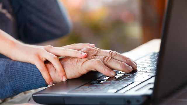 A young caregiver helps an elderly male relative use a computer by guiding his hand.