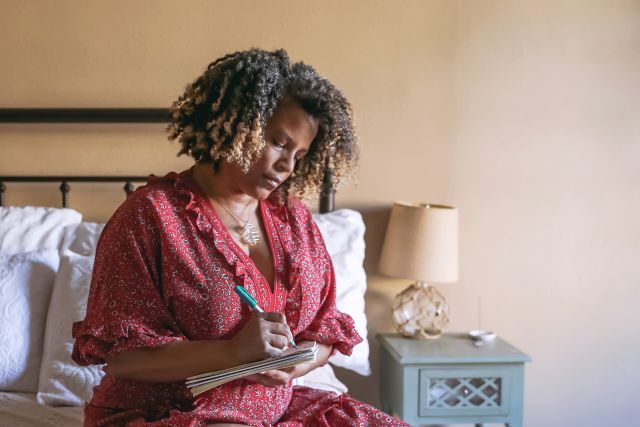 a middle aged Black woman sits on the edge of her bed and writes in her journal