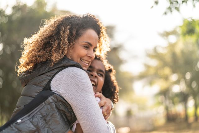 Black woman hugs child outside on sunny day near trees