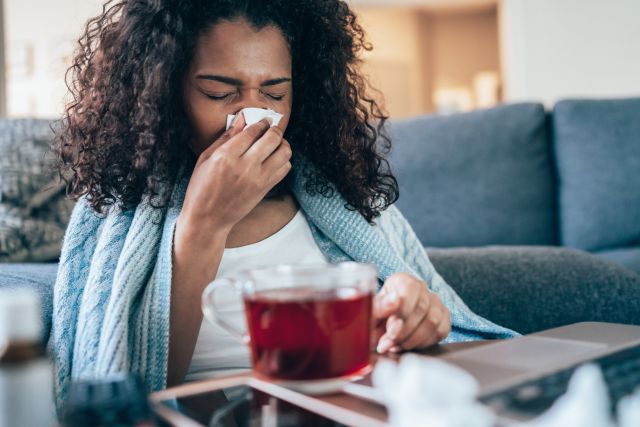 Black woman home sick with a cold or the flu, blowing her nose while sitting on the sofa
