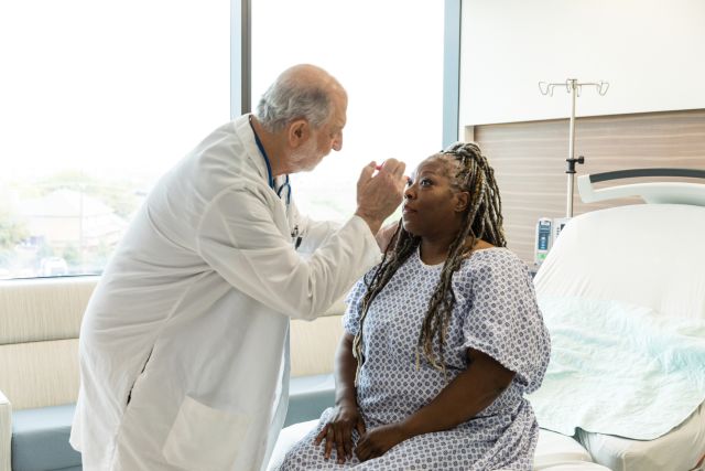 a middle aged Black woman is examined by an older white male doctor in a hospital room; the doctor shines a light in the patient's eyes to check for signs of a concussion