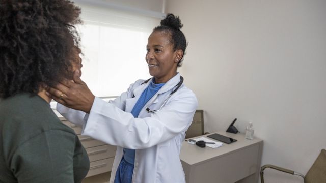 A Black healthcare provider examines a female patient's thyroid gland during an appointment.