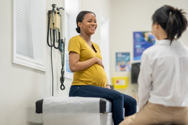 a young pregnant Black woman wearing a yellow shirt sits on an examination chair and smiles as she speaks with her Asian American female healthcare provider about getting vaccinated for COVID  