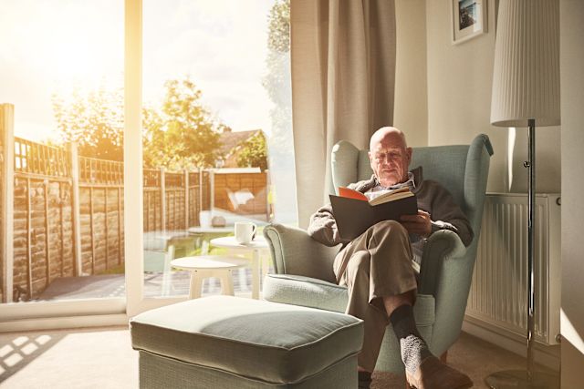 An older white adult man sits in his chair reading as natural sunlight floods into the room from a glass sliding door next to him.