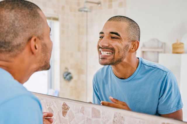 A young Black man stands in front of his bathroom mirror looking closely at his teeth to measure his progress improving his oral health through regular brushing and flossing