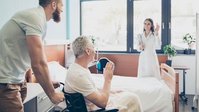 A patient waves while being transported in a wheelchair with nurses standing by.