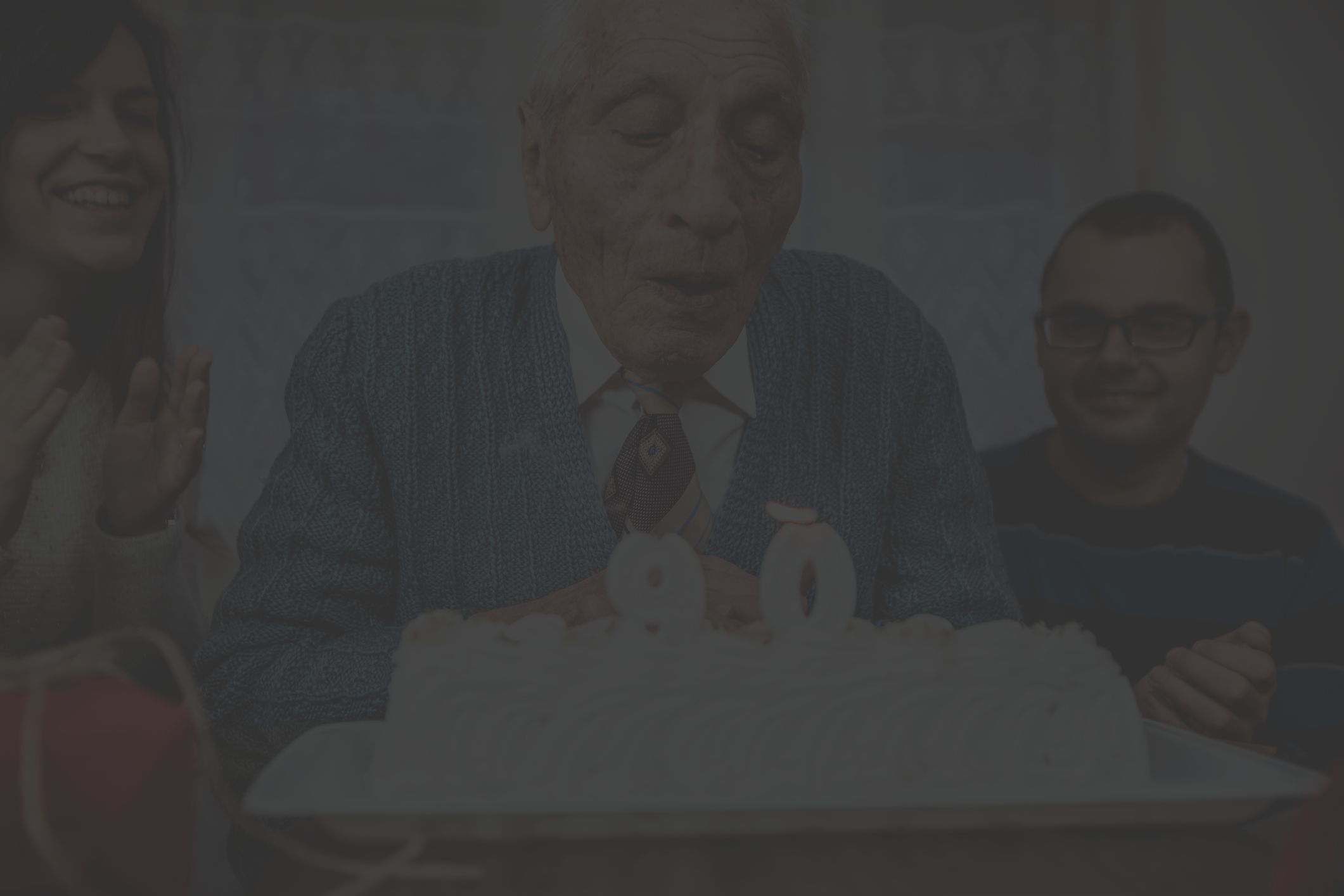 an older white man celebrates his 90th birthday with loved ones; he sits in front of a birthday cake and blows out the candles