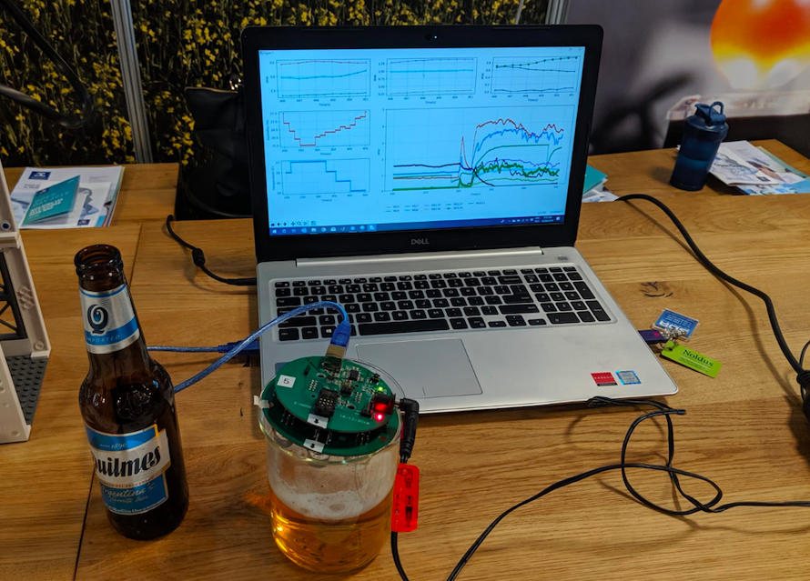 The e-nose device looks like a microchip. It's covering a glass of beer and plugged into a laptop via USB. The laptop shows a range of data, and a beer bottle sits next to the glass on a wood desk. 