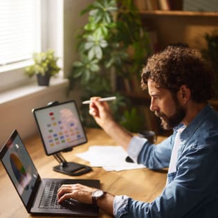A man with curly hair and a beard works on a laptop and a tablet with a stylus at a wooden desk.