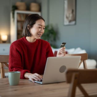 A woman in a red sweater holds a credit card while using a laptop at a wooden table.