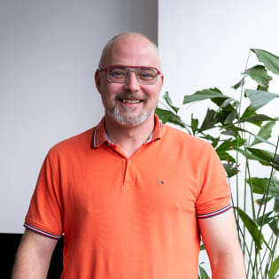 A smiling man with a beard and glasses wearing an orange polo shirt.