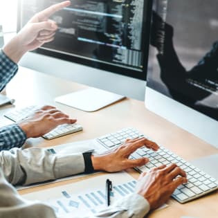 Two people coding on computers, one pointing at a screen displaying lines of code.