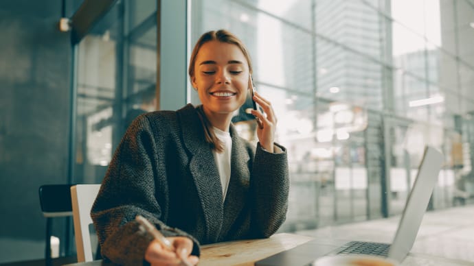 A smiling woman in a dark blazer talks on her smartphone while writing at a table with a laptop.